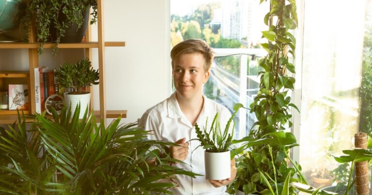 Lainey Martin stares off-camera, surrounded by houseplants, smiling slightly as she holds a small potted plant.