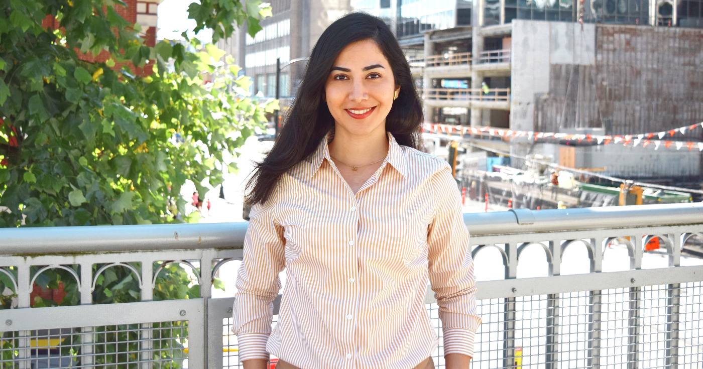 Ghazaleh stands on a sidewalk with a metal grate railing behind her, looking straight at the camera, with a green bush behind her to her right and a parkade to her left.