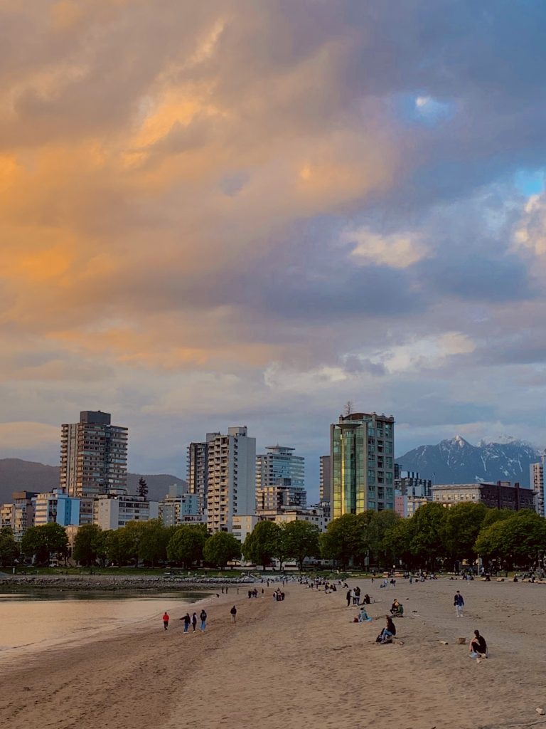 A view of English Bay in Vancouver, British Columbia at sunset with beach in the foreground and apartment buildings and the Coast mountain range in the background.