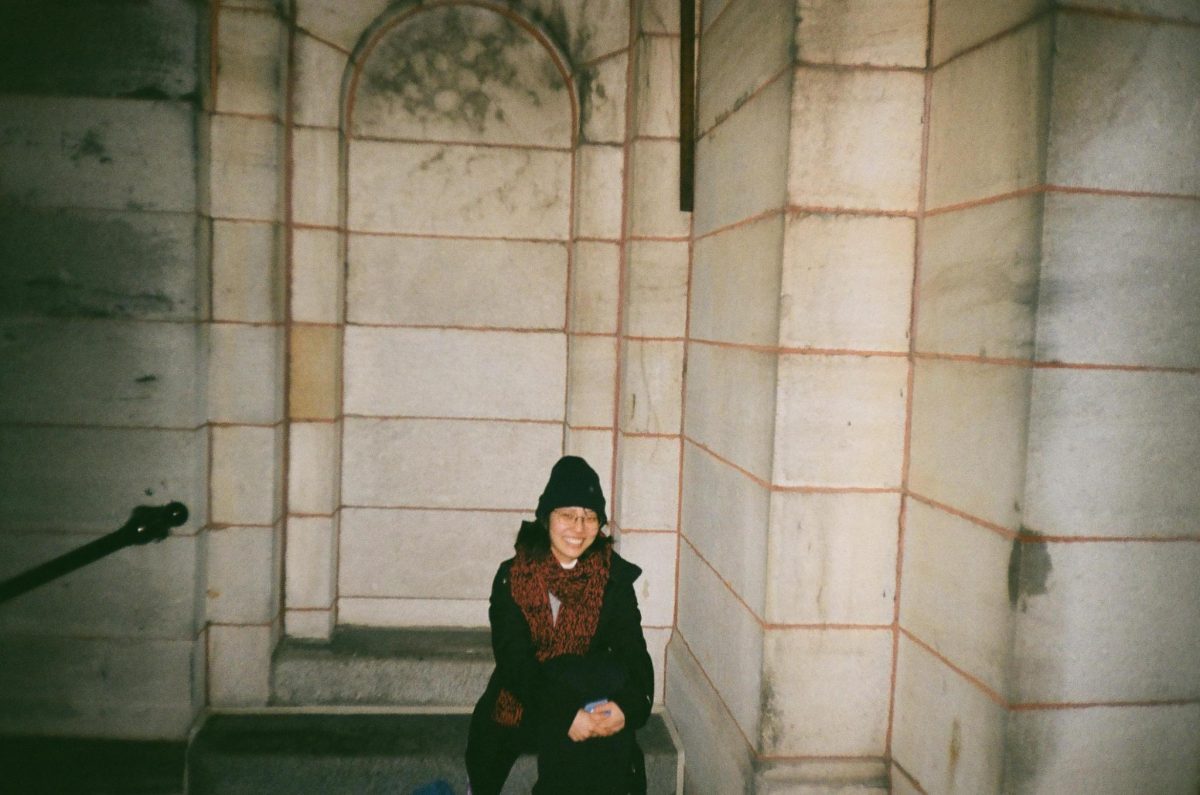 Robyn Lee in a black coat and toque, sitting on a stone bench in a stone building.