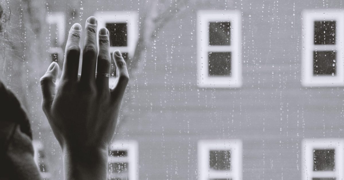 A woman presses her right hand up to a window with rain droplets running down it while across from her is an apartment building with white windowpanes.