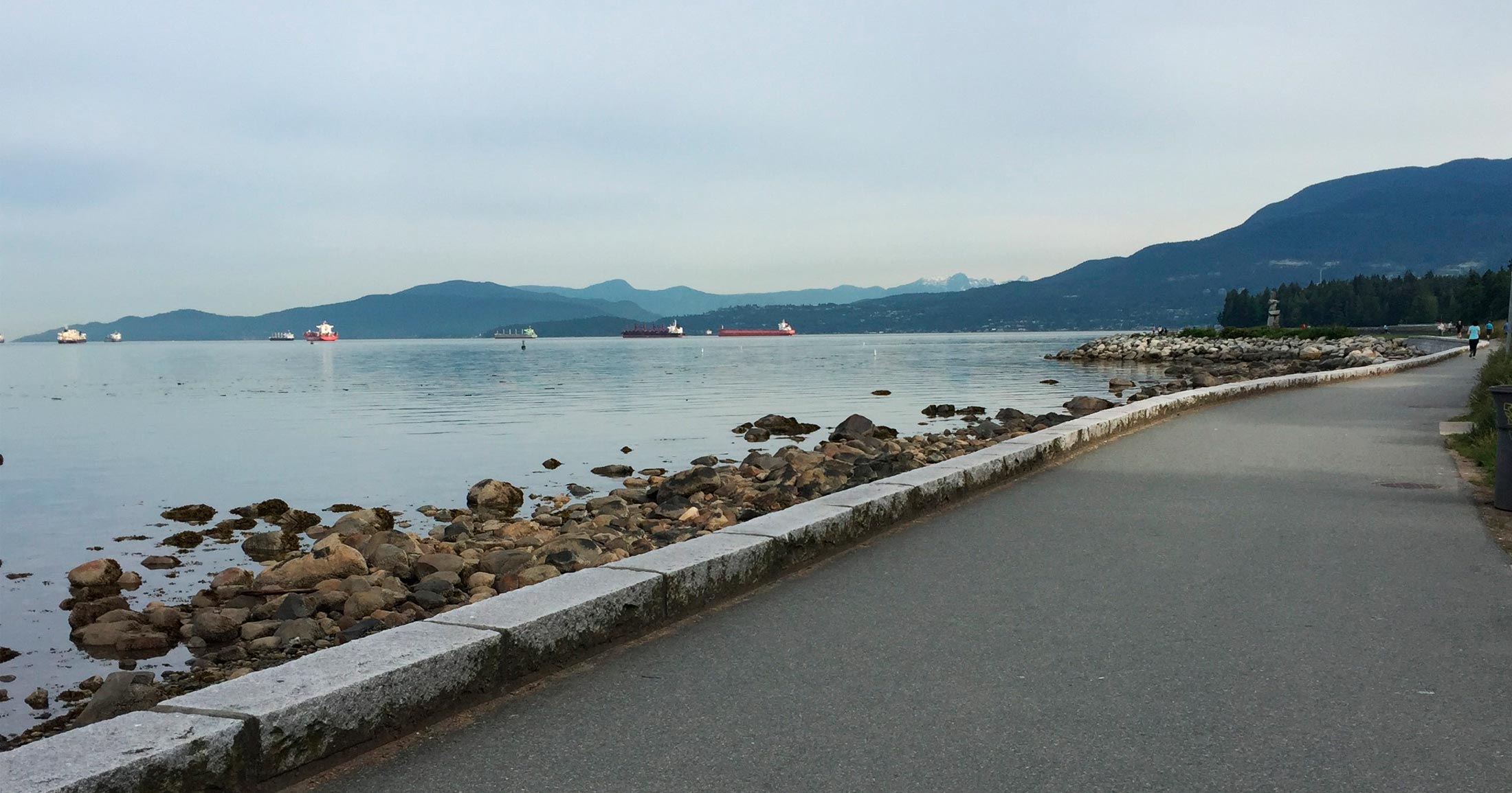 A pathway runs along the waters of the Vancouver coastline toward the mountains and grey clouded sky.