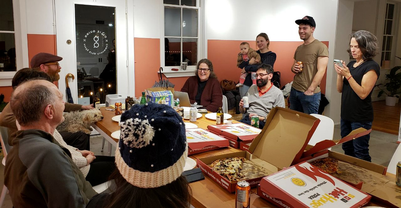 A group of residents sit and stand around a table with pizzas laying on top.