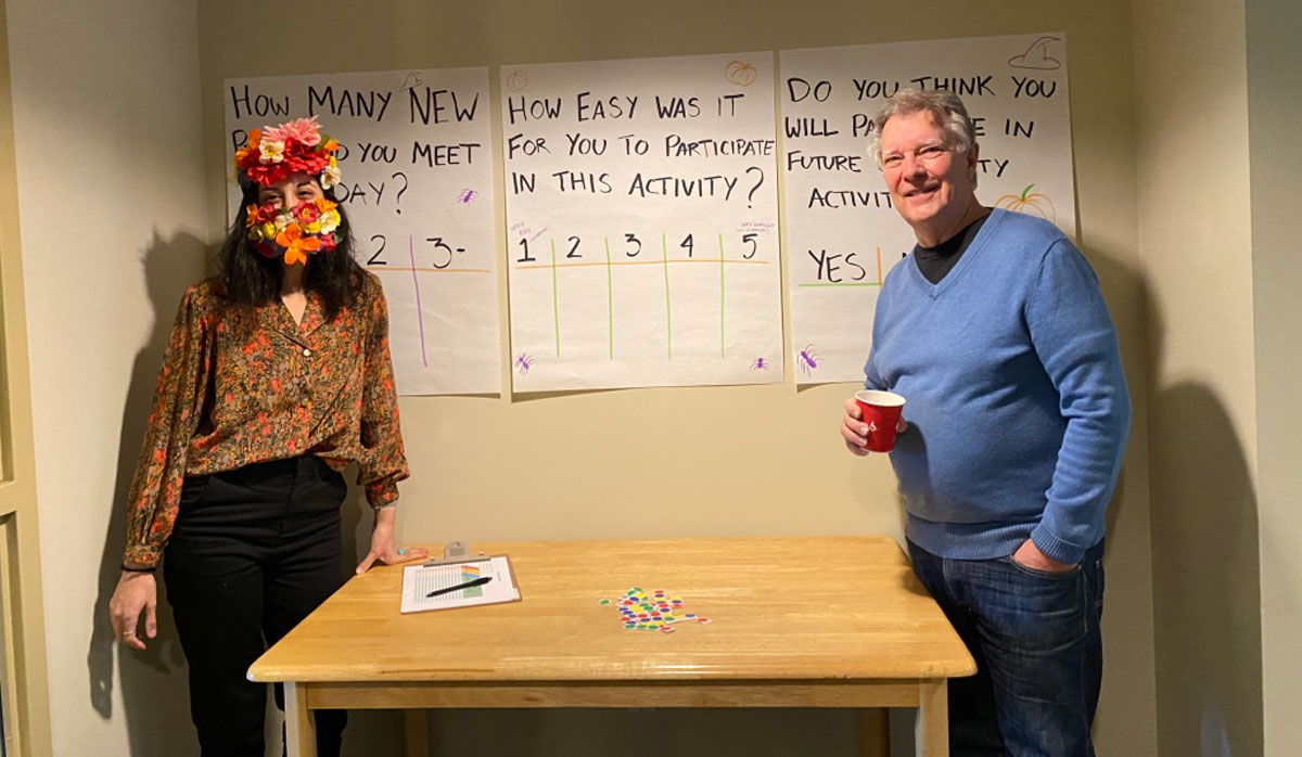 Two community connectors stand in a room with a table and paper taped up on the wall with titles and survey questions.