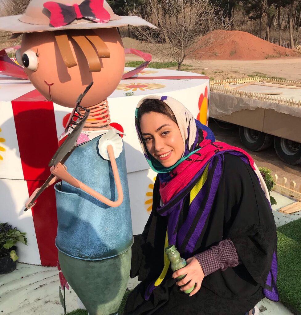 Niloofar wears a colourful headscarf while holding a bottle of green paint, kneeling next to a sculpture that she helped design in Mashhad, Iran.