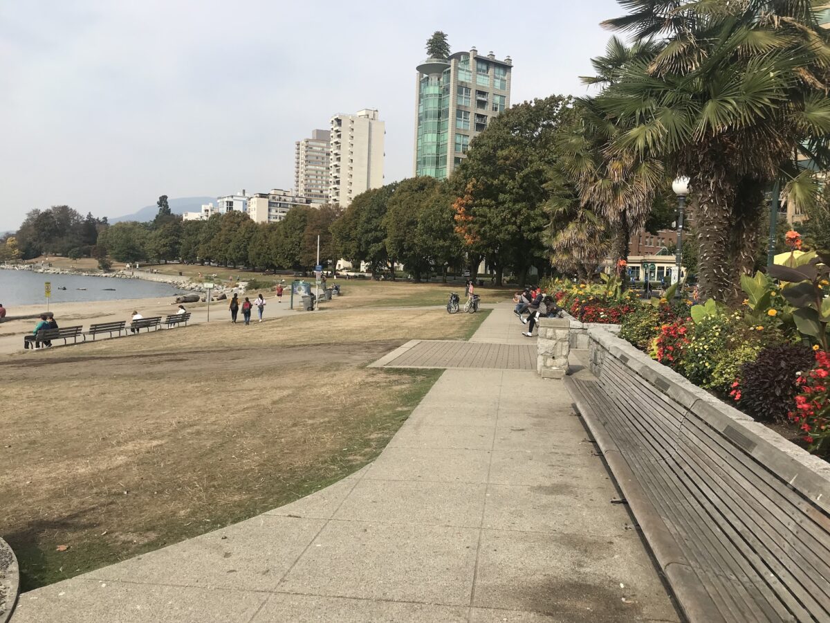 The public walkway along English Bay in downtown Vancouver, BC with benches for sitting, lush foliage and trees, and tall buildings in the background.