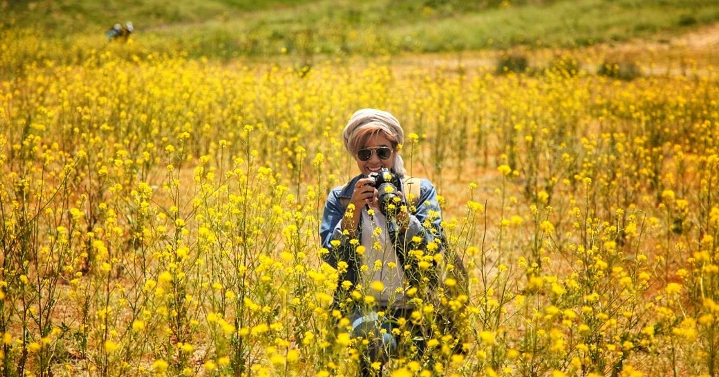 Sogol Haji Hoseini stands in a field of yellow, wildflowers wearing sunglasses while smiling and holding a camera.