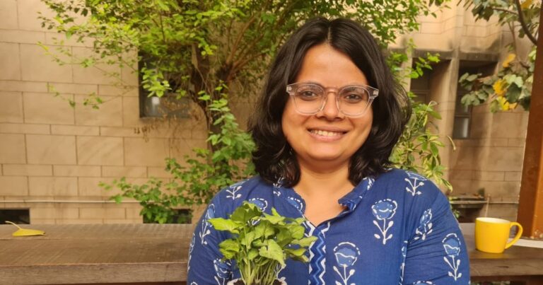 Sreya Ajay smiles, wearing clear framed glasses and a blue shirt with white flowers while holding a potted plant and stands in front of a tree-lined concrete building.