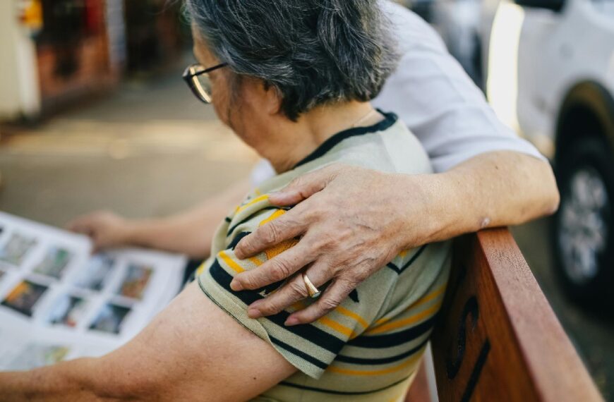 Two older persons sit on a bench reading the newspaper with one person's left arm over her shoulder.