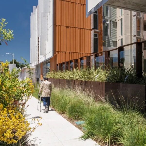 An older adult walks with the aid of a walker along a grass and tree-lined path in front of Lakeside Senior Apartments in Oakland.