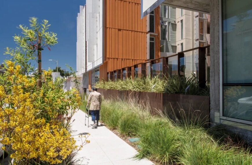An older adult walks with the aid of a walker along a grass and tree-lined path in front of Lakeside Senior Apartments in Oakland.