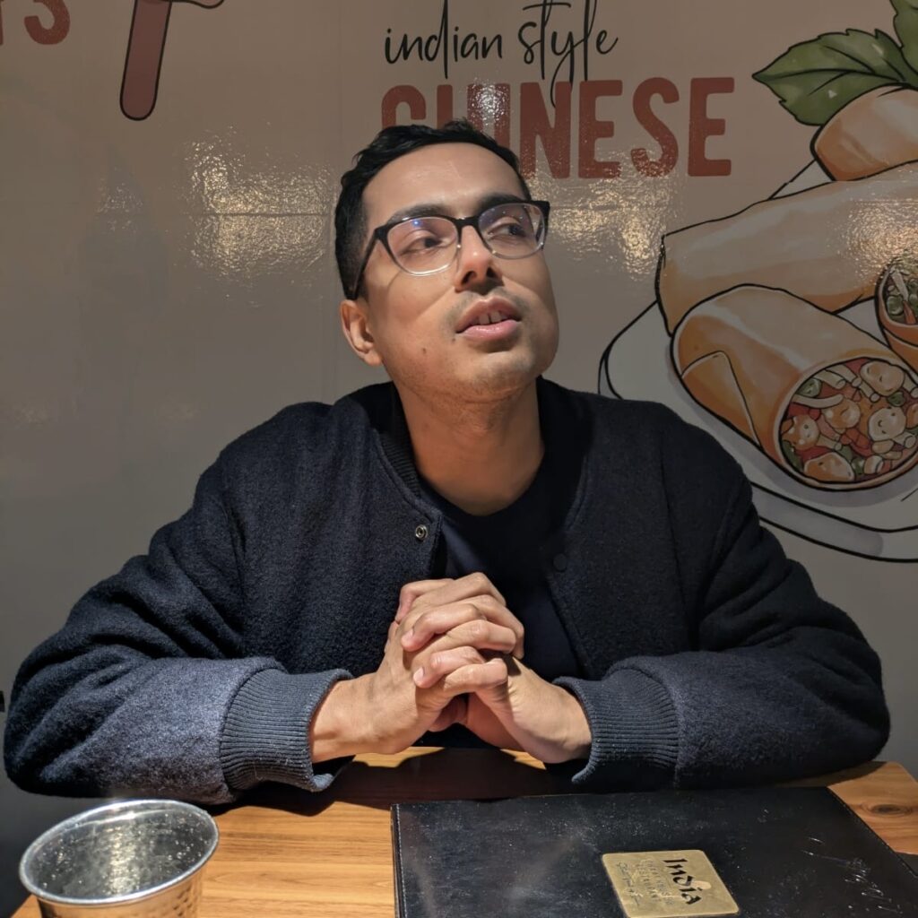 Kishore Seetharaman sits at a table at an Indian restaurant with his hands clasped and his gaze off to one side.
