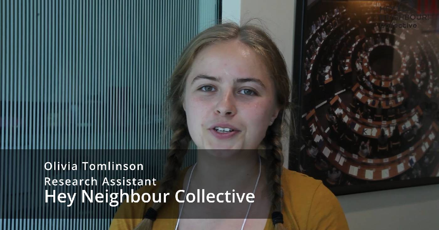 Olivia Tomlinson looks at the camera while being interviewed in this still excerpt from a video. Text is overlaid with her name and her title, "Research Assistant, Hey Neighbour Collective."