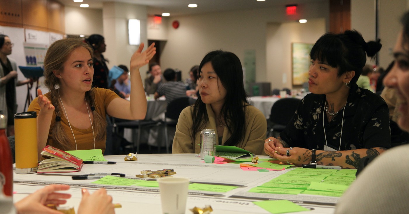 Olivia speaks with her arms raised at a table during the Living Together Symposium in May 2025.
