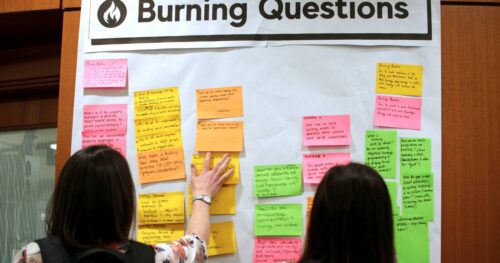 Participants at the Living Together 2025 Symposium place colourful Post-It Notes on a wall sheet titled, "Burning Questions."