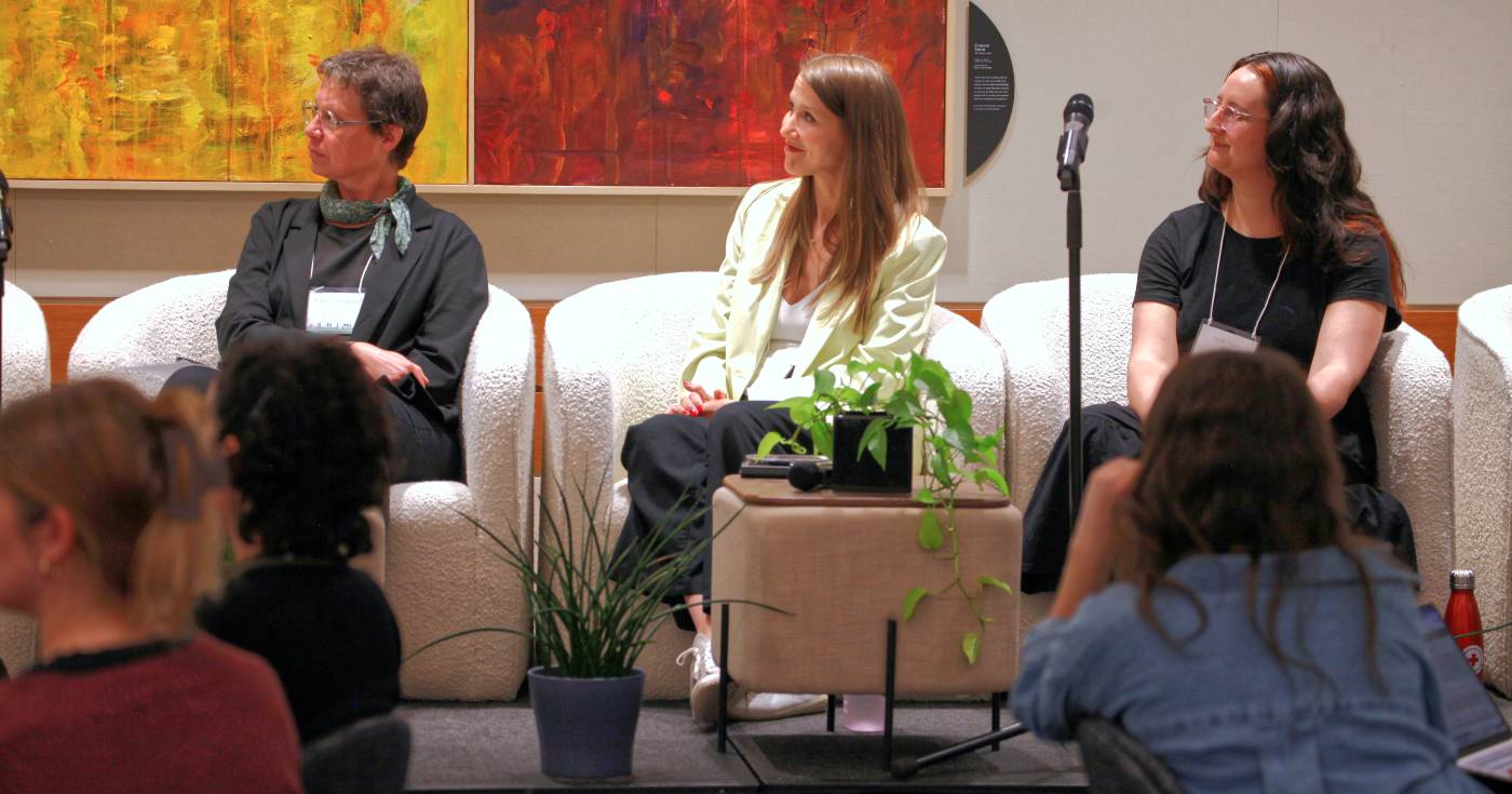 Panelists at the Living Together Symposium sit in white couches against a yellow and red artwork, with green plants and participants in the foreground.