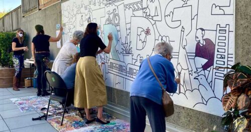 A group of people create a mural outside along a sidewalk.